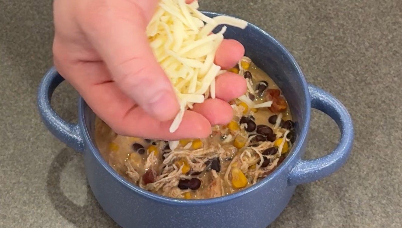 Hand sprinkling shredded cheese over a blue ceramic bowl of creamy chicken chili with black beans, corn, and shredded chicken