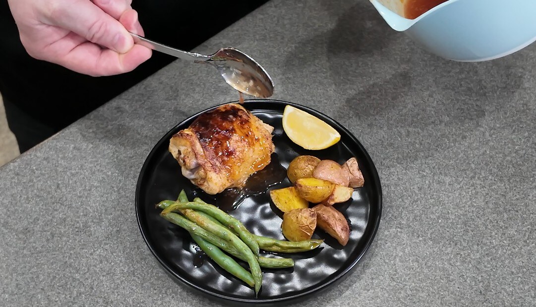Spooning hot honey over a roasted chicken thigh with potatoes, green beans, and lemon